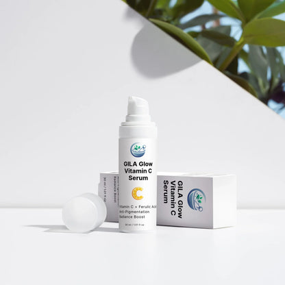 An image of the GILA Glow Vitamin C Serum with lid off, and the box on its side. It's on a white outdoor background, with shrubs peeking up from behind
