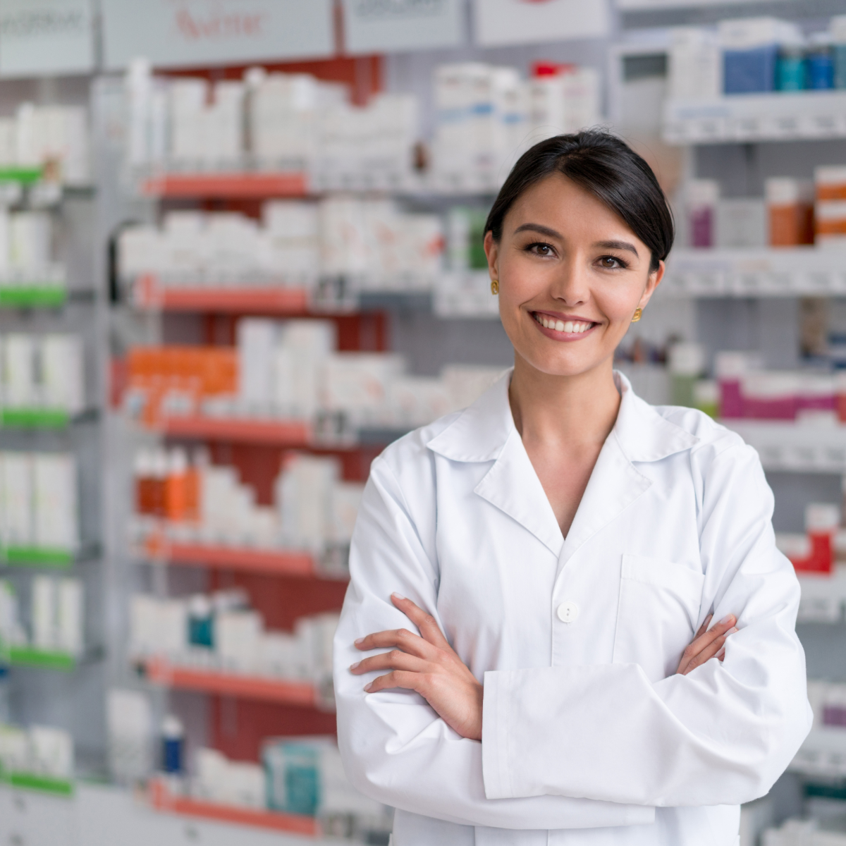 A photo showing a beautiful female pharmacist wearing a lab coat and posing in front of a display of medications